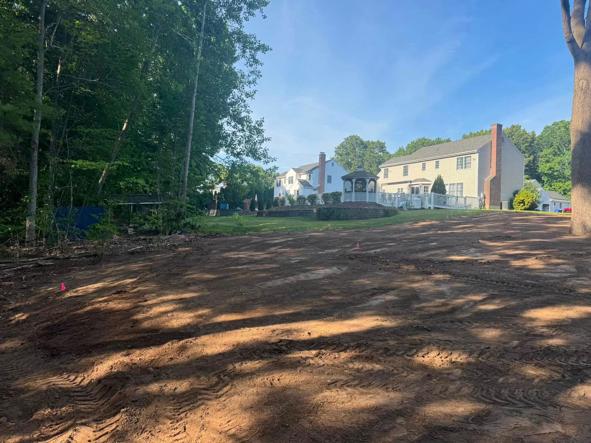 Cleared dirt lot beside trees and houses under a bright blue sky