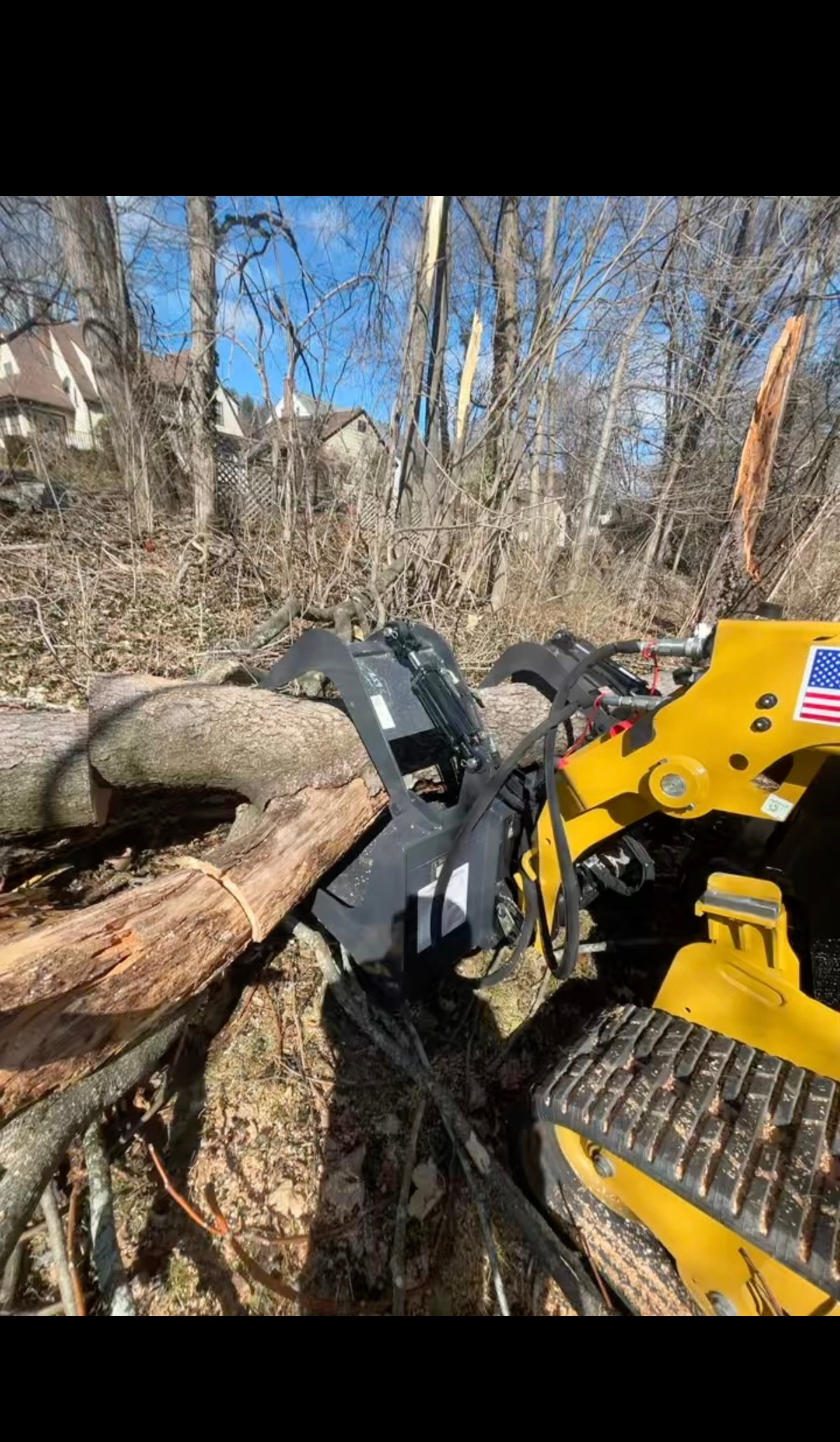 Yellow skid steer lifting a log in a leafless wooded area