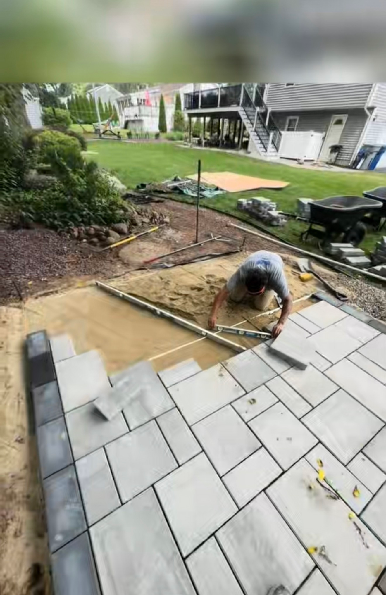 Worker laying gray pavers on a backyard patio beside a grassy lawn and house.