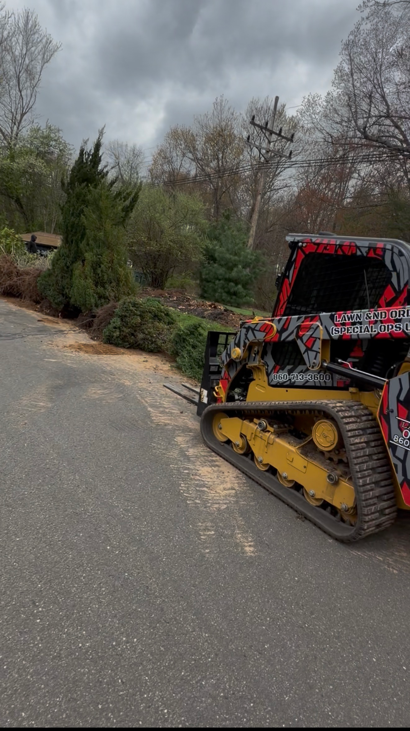 Tracked snow blower clearing a wet driveway beside trees under a cloudy sky