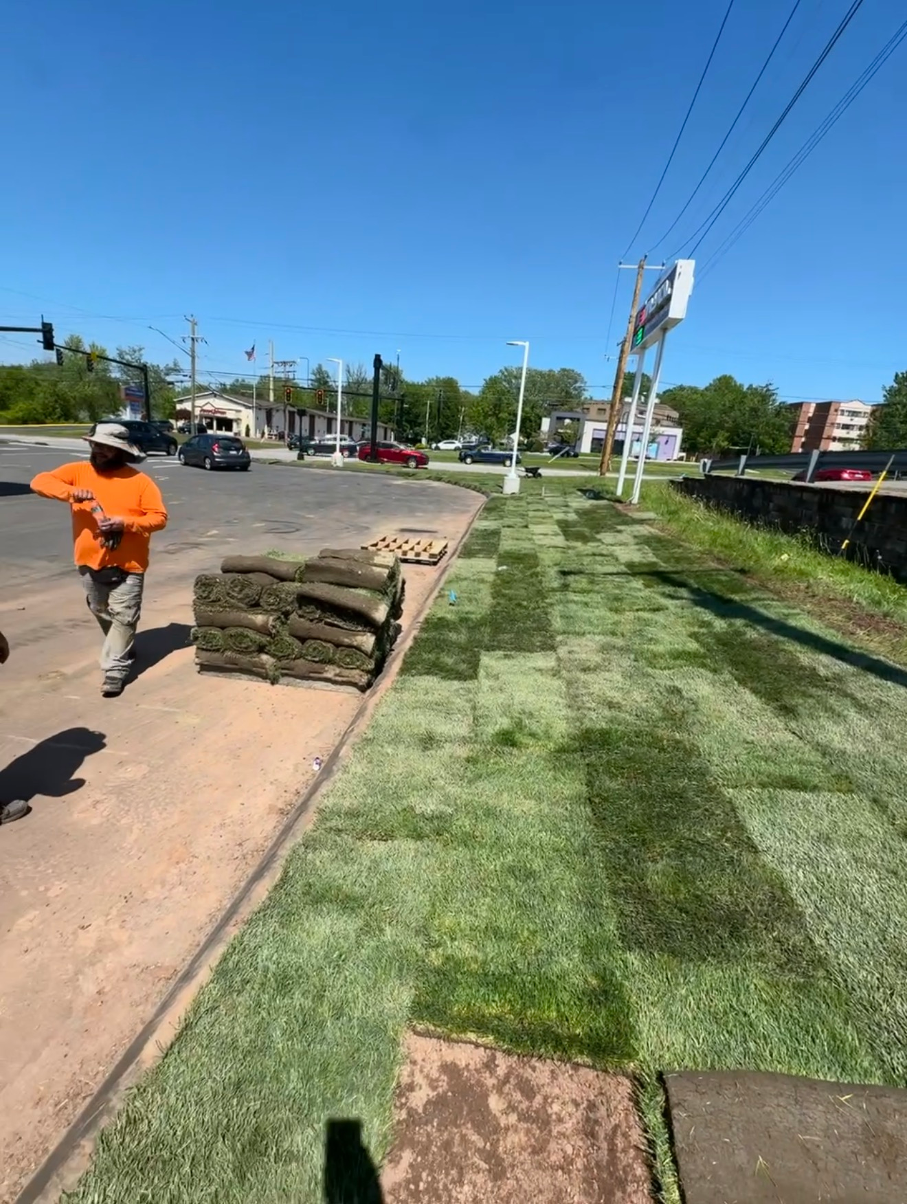 Worker carrying grass sod along a roadside sidewalk under a bright blue sky