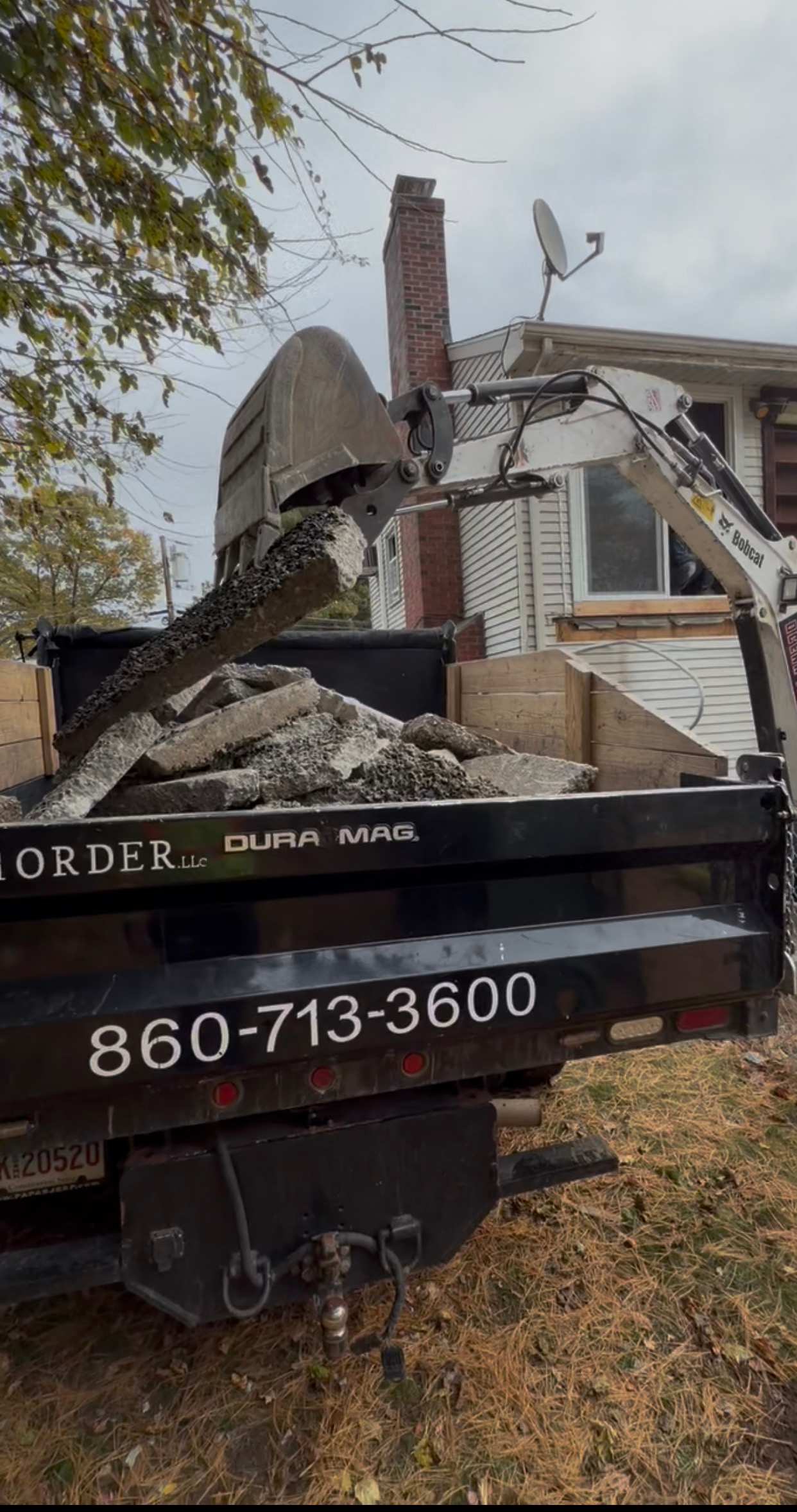 Excavator demolishing a brick house beside a truck with phone number 860-713-3600.