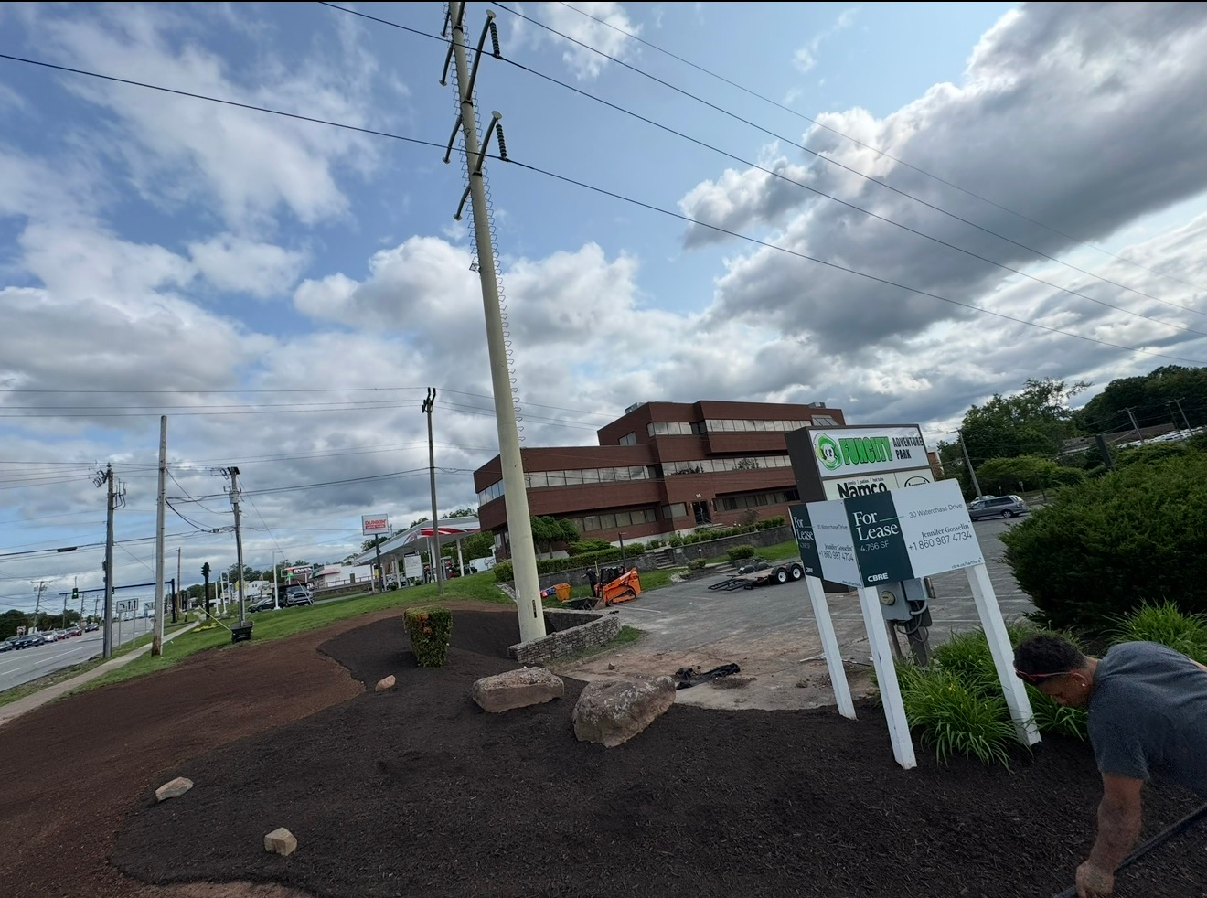 Roadside construction area with utility poles, cloudy sky, and a sign by a building; worker at right.