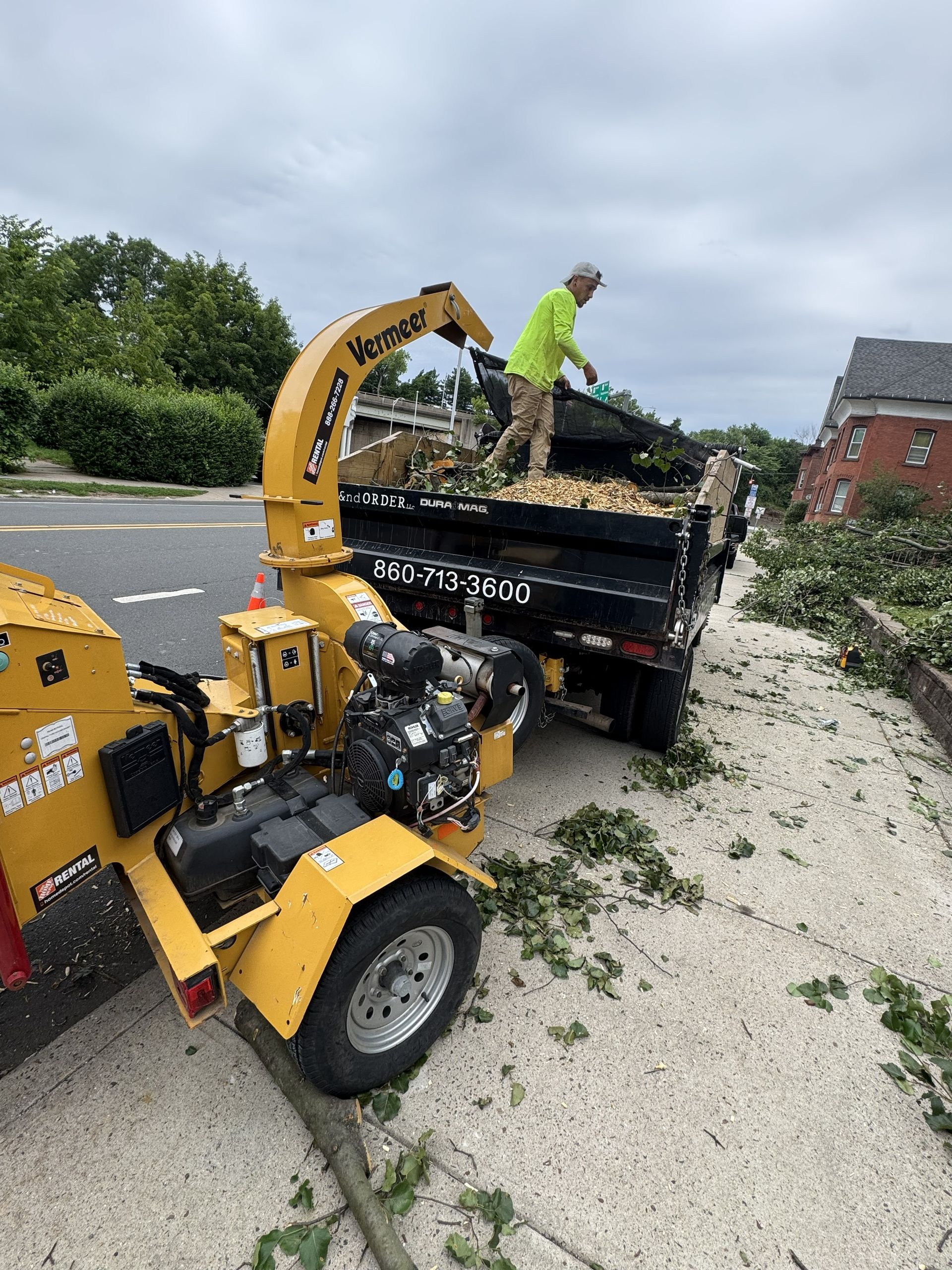 Worker loading tree debris into a chipper beside a truck on a roadside.