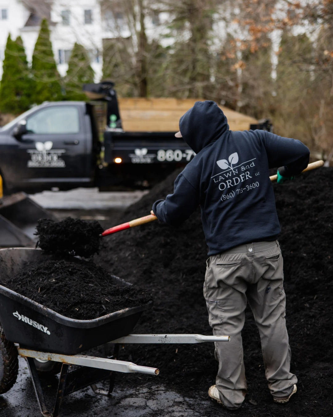 Worker shoveling dark mulch beside a truck in a yard, wearing a hooded sweatshirt and gloves