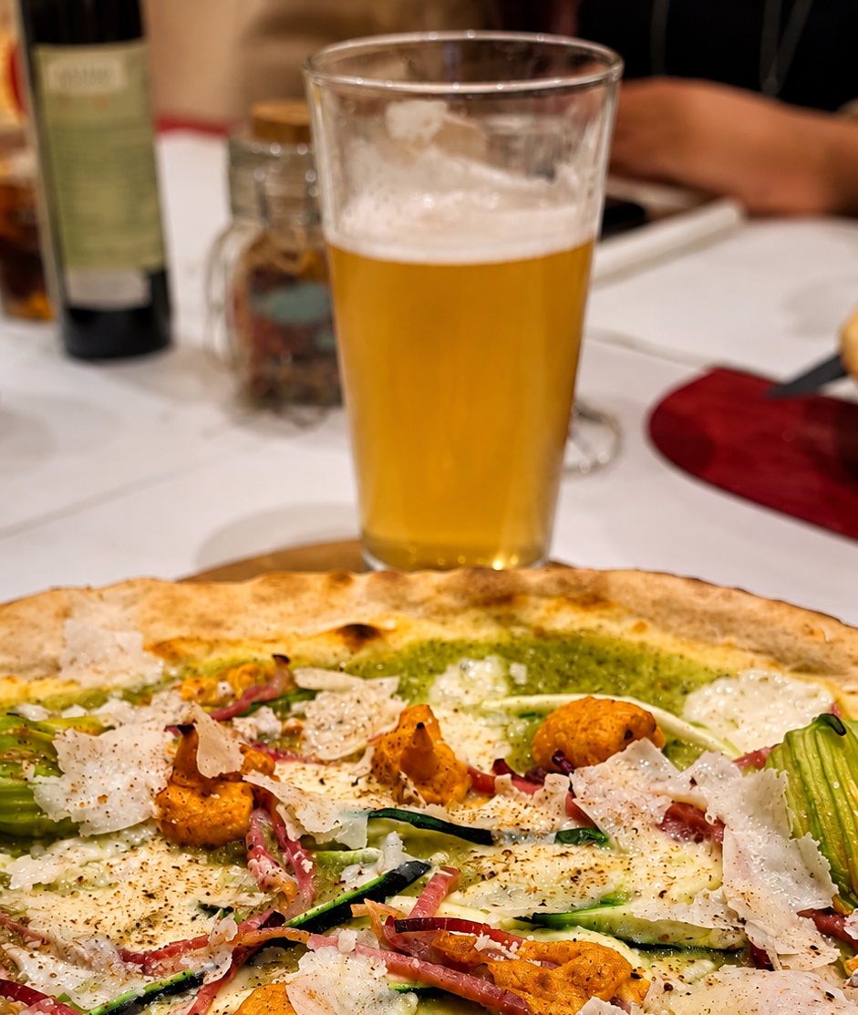 Una pizza e un bicchiere di birra mezzo pieno su un tavolo di ristorante, con piatti e vino sullo sfondo.