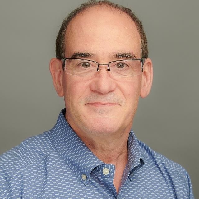 Man with glasses and a blue shirt smiles at the camera against a gray background.