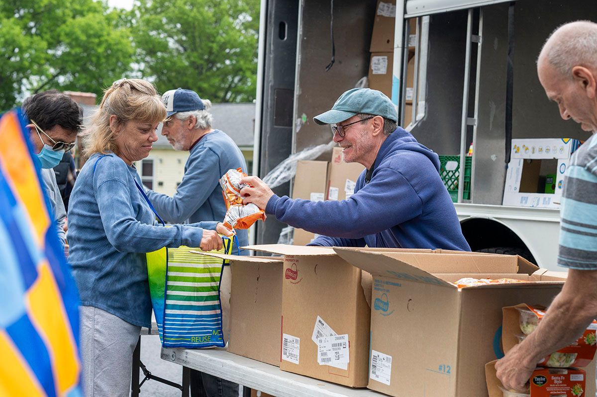 People distributing food from a truck to others outdoors; boxes and a table are present.