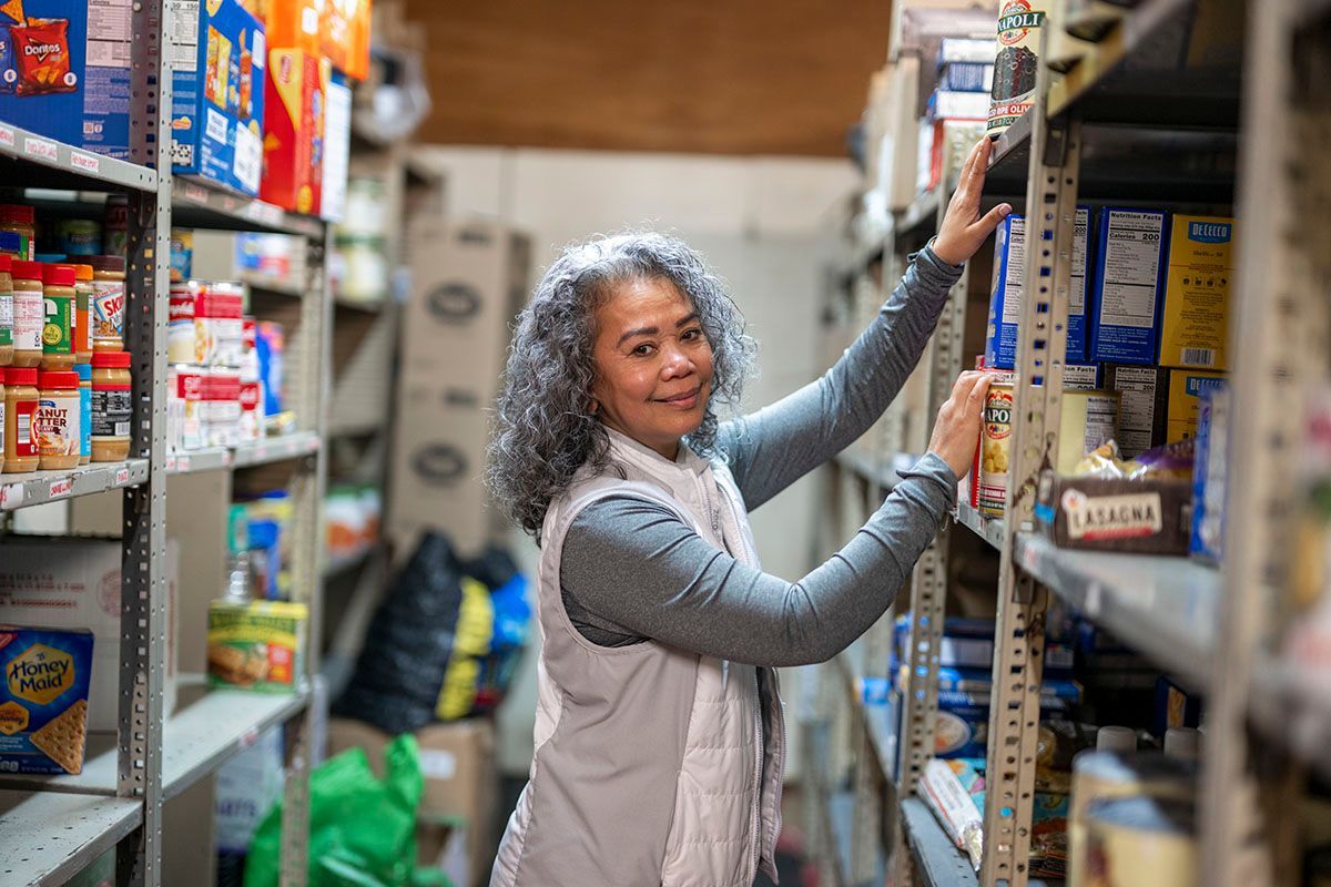 Woman reaching for items on a shelf in a pantry, smiling. Shelves filled with packaged food items.