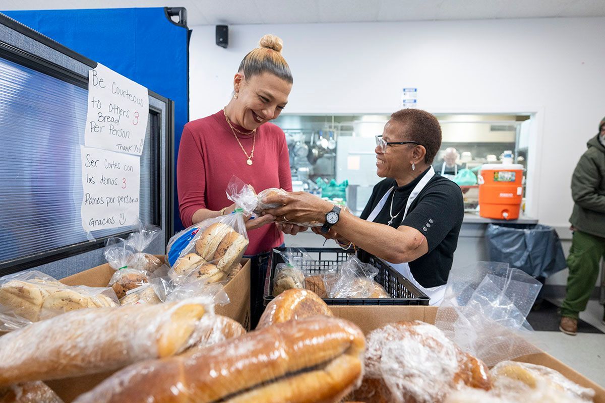 Woman receives bread from another woman at a food bank, smiling. Boxes of bread are in front of them.