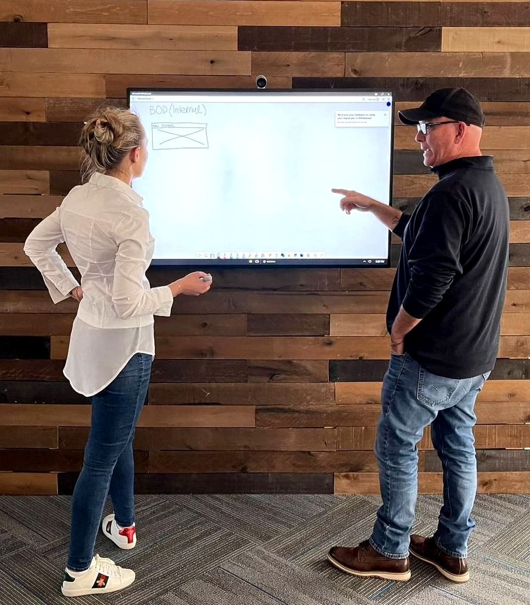 Nicole and Jeff looking at a screen on a wooden wall, man points.