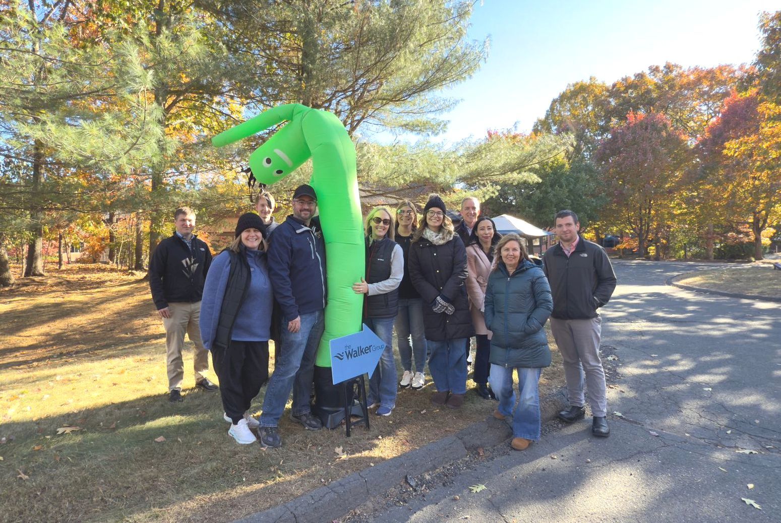 Group of people pose with a tall, green inflatable figure outdoors in front of trees. at the Ewaste event at The Walker Group. 