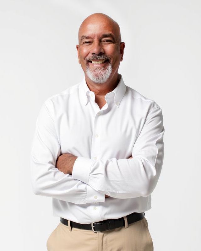 Man with graying hair and beard smiles, arms crossed, wearing a white button-down shirt and khakis.