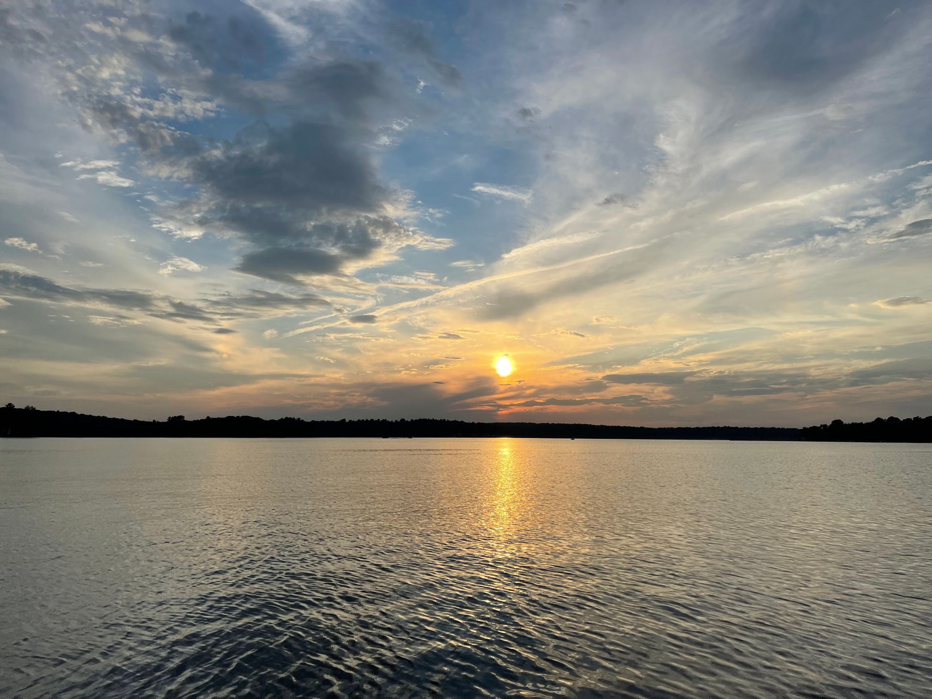 Sunset over a calm lake, with golden sunlight reflecting on the water and a cloudy blue sky.