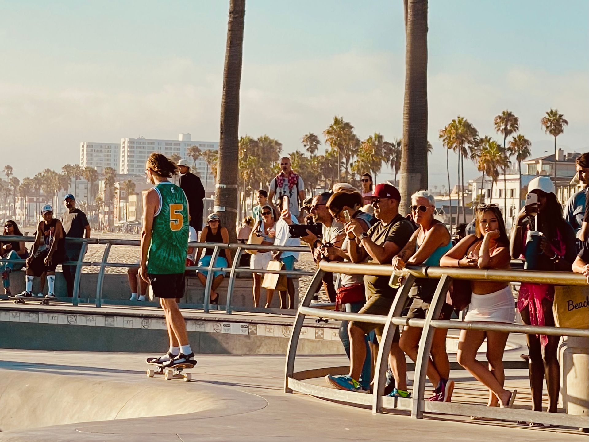 Skateboarder in a green jersey performs near a crowd in a sunny skatepark. Palm trees and buildings in background.