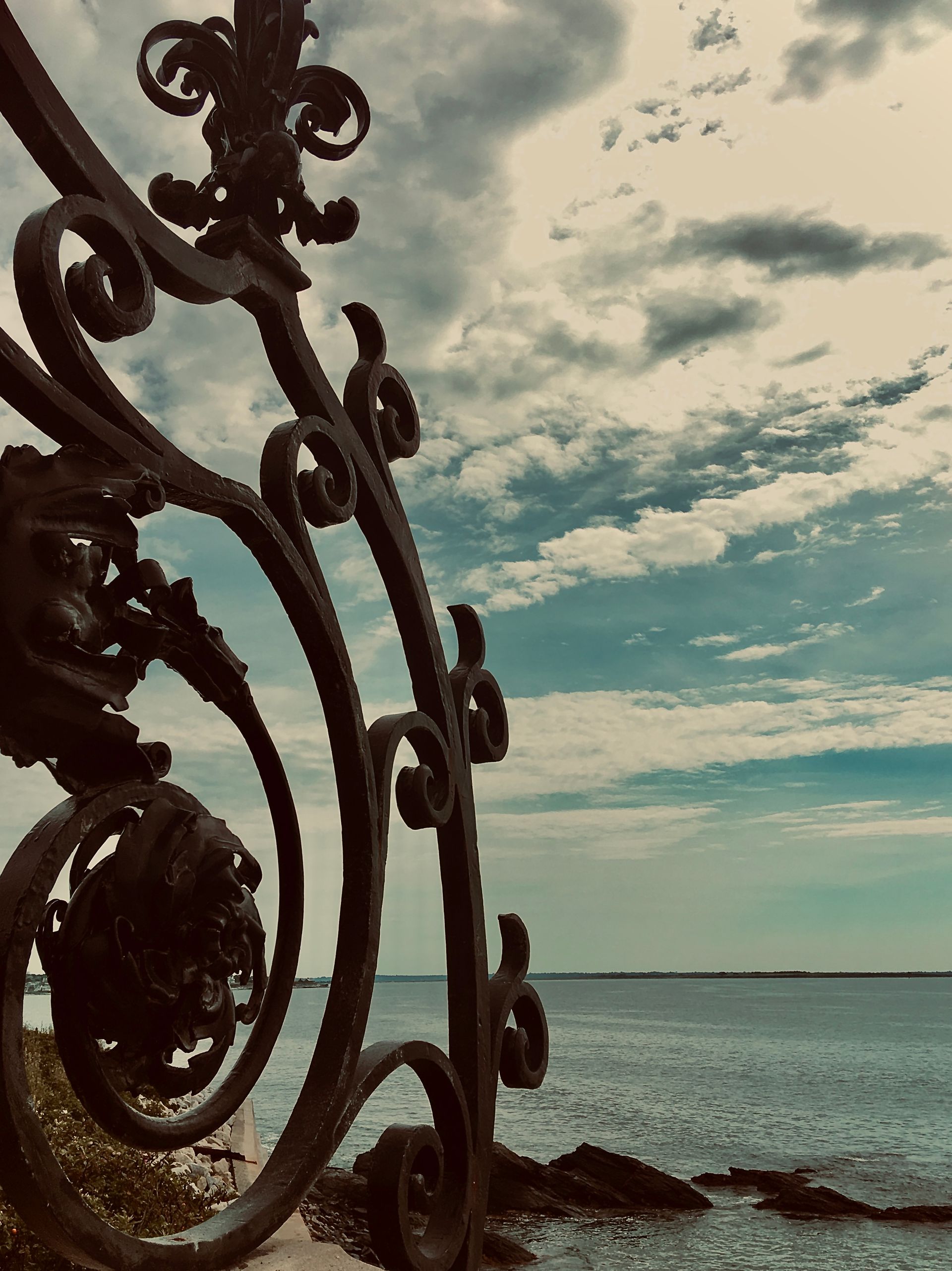 Ornate, dark metal gate framing a seascape with cloudy sky.