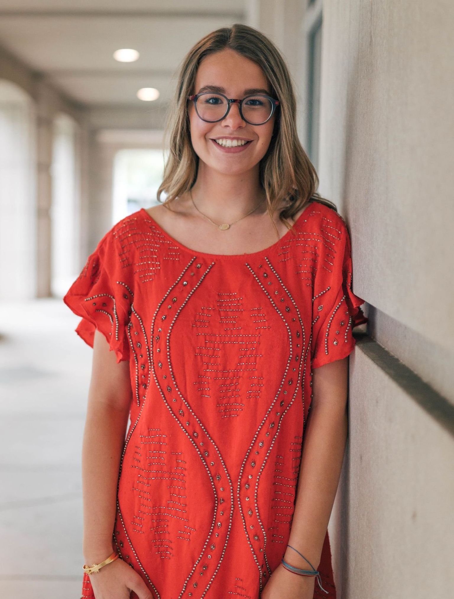 Woman with glasses smiles, wearing red dress, leans against a beige wall in a covered walkway.