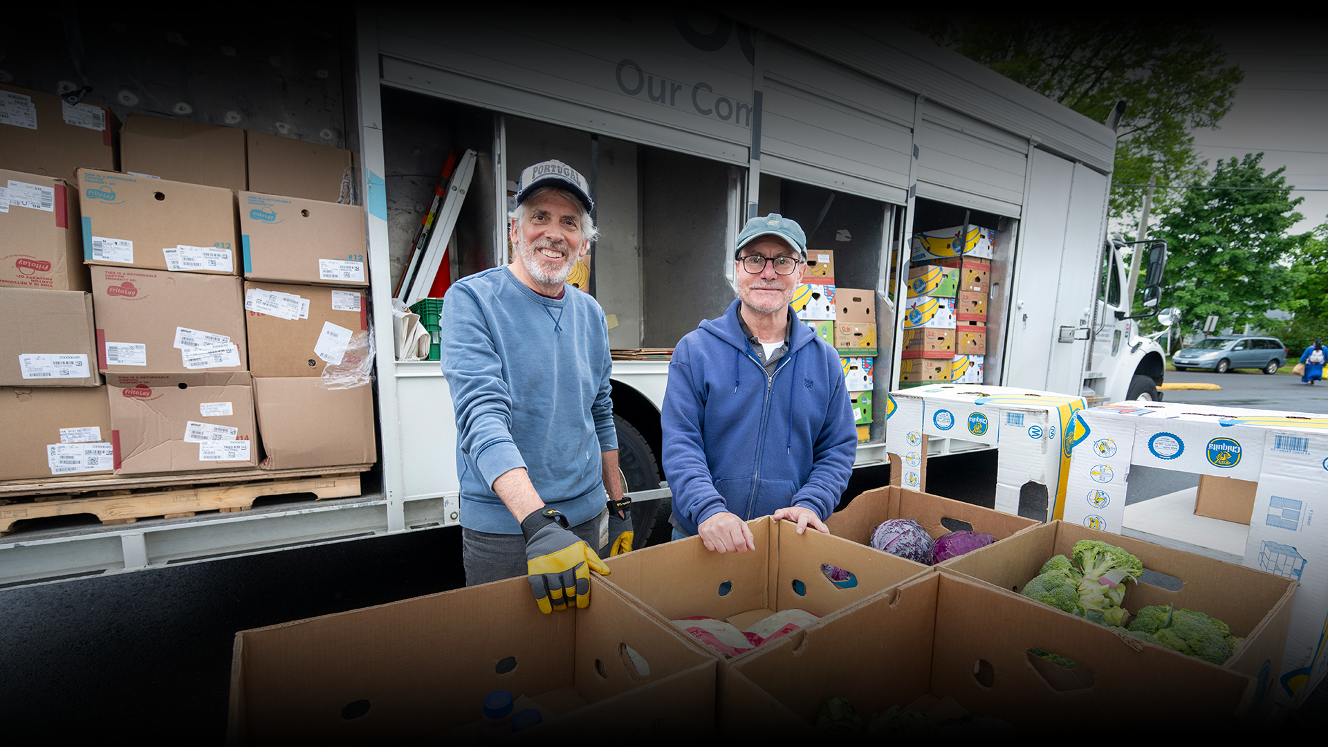 Two men smiling, sorting produce from truck boxes in parking lot, boxes and boxes of food visible.