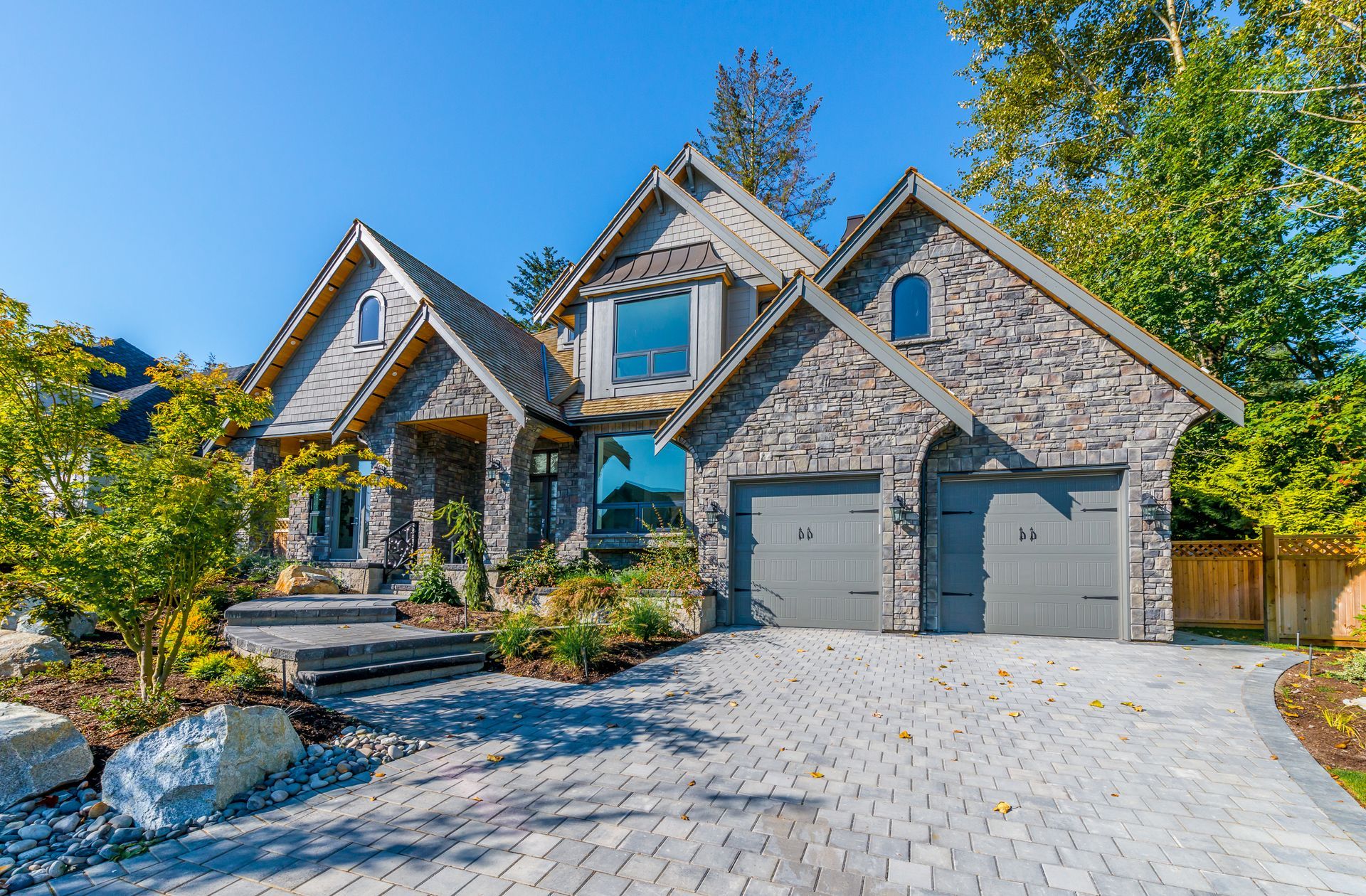 Stone-clad house with a gray paved driveway, two-car garage, and landscaped yard under a bright blue sky.