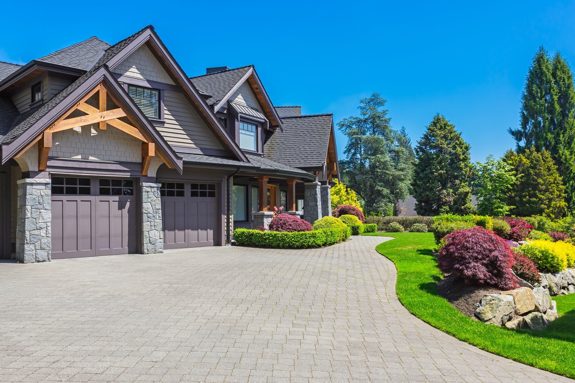 Stone-faced house with a two-car garage, manicured lawn, and landscaped gardens under a blue sky.
