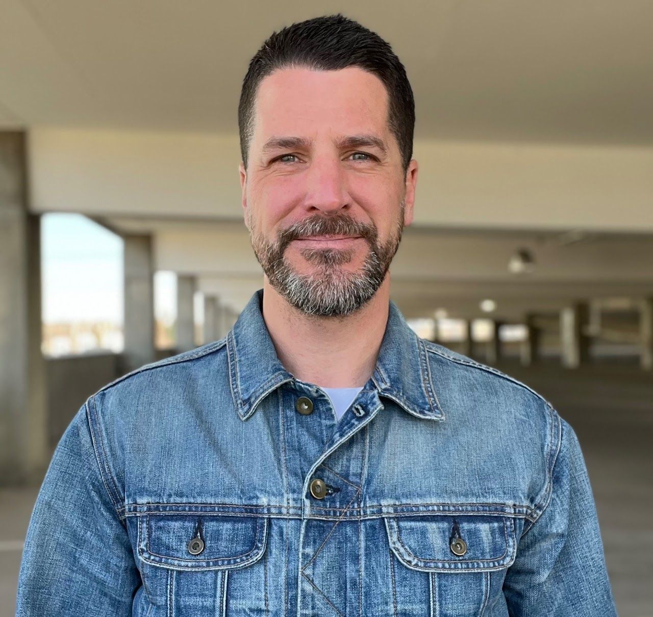 Man in a denim jacket smiling indoors with a blurred hallway background
