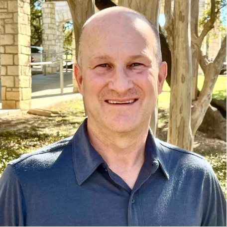 Smiling bald man in a blue shirt standing outdoors near stone buildings and trees