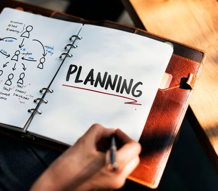 Notebook with “PLANNING” written on a page, beside a hand holding a pen and a leather folder.