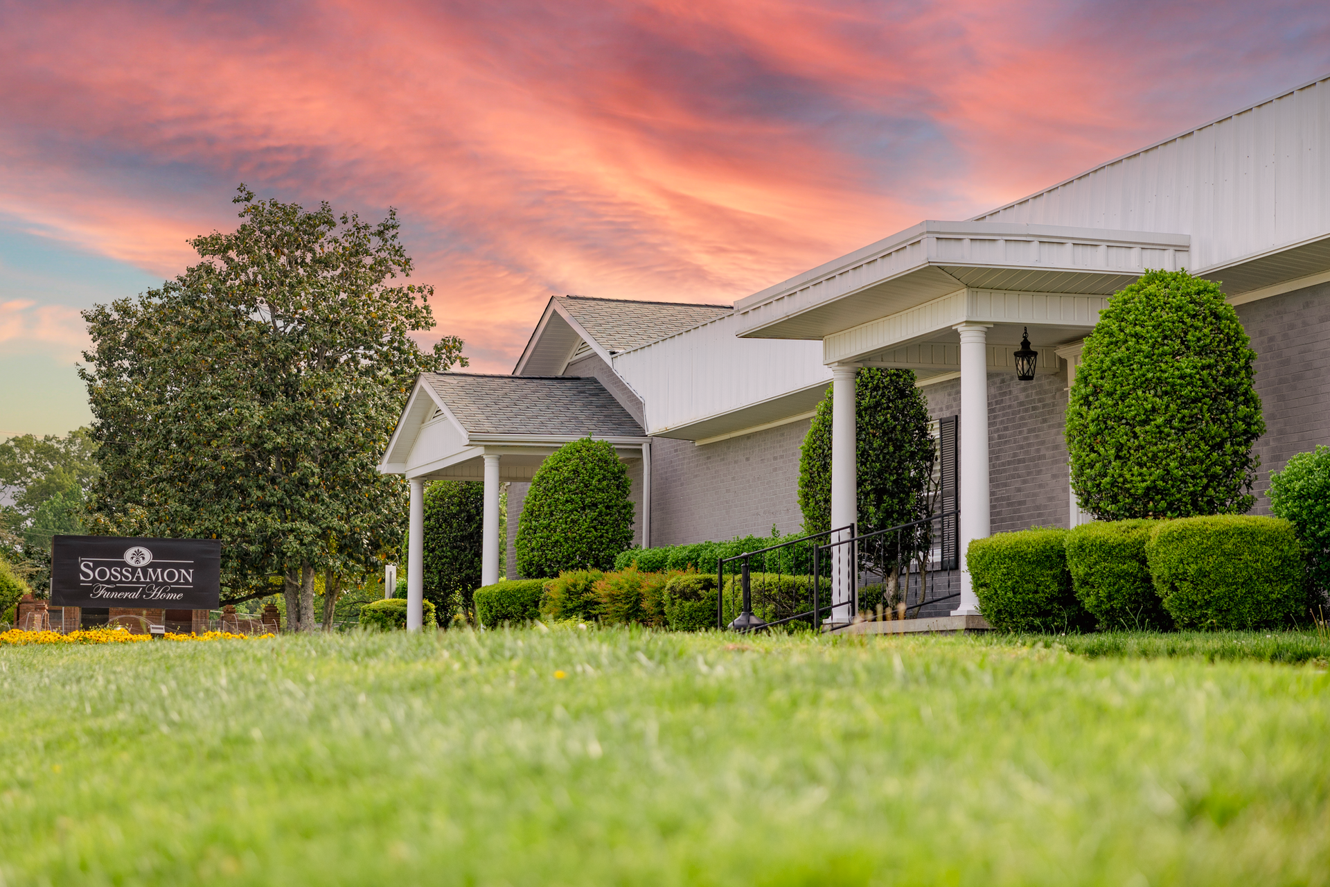 A long, single-story building with a white exterior, columns, and trimmed hedges under a pink and orange sunset sky.
