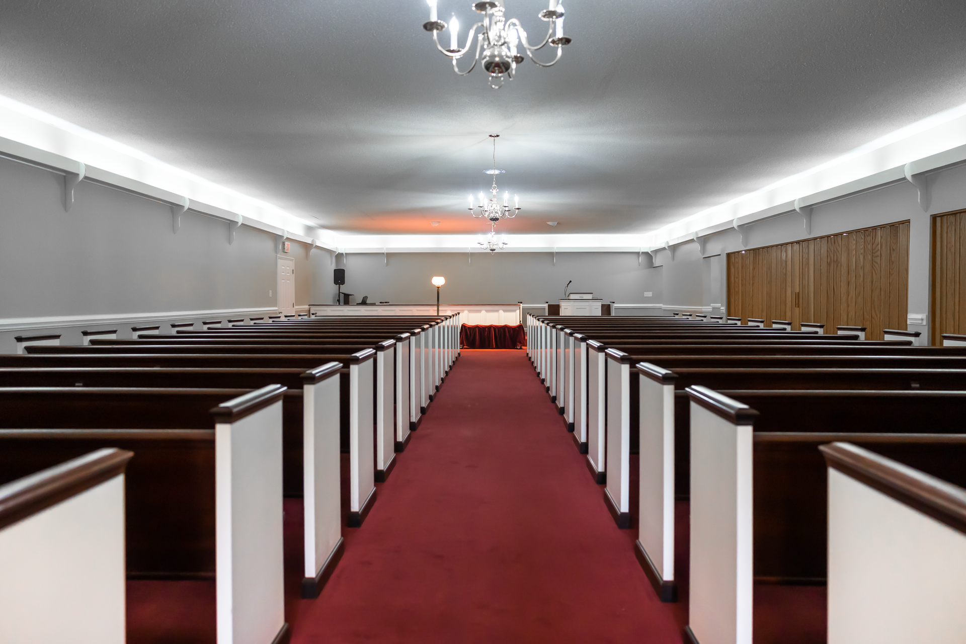 A long, empty church sanctuary with rows of wooden pews, a red carpeted aisle, and hanging chandeliers.