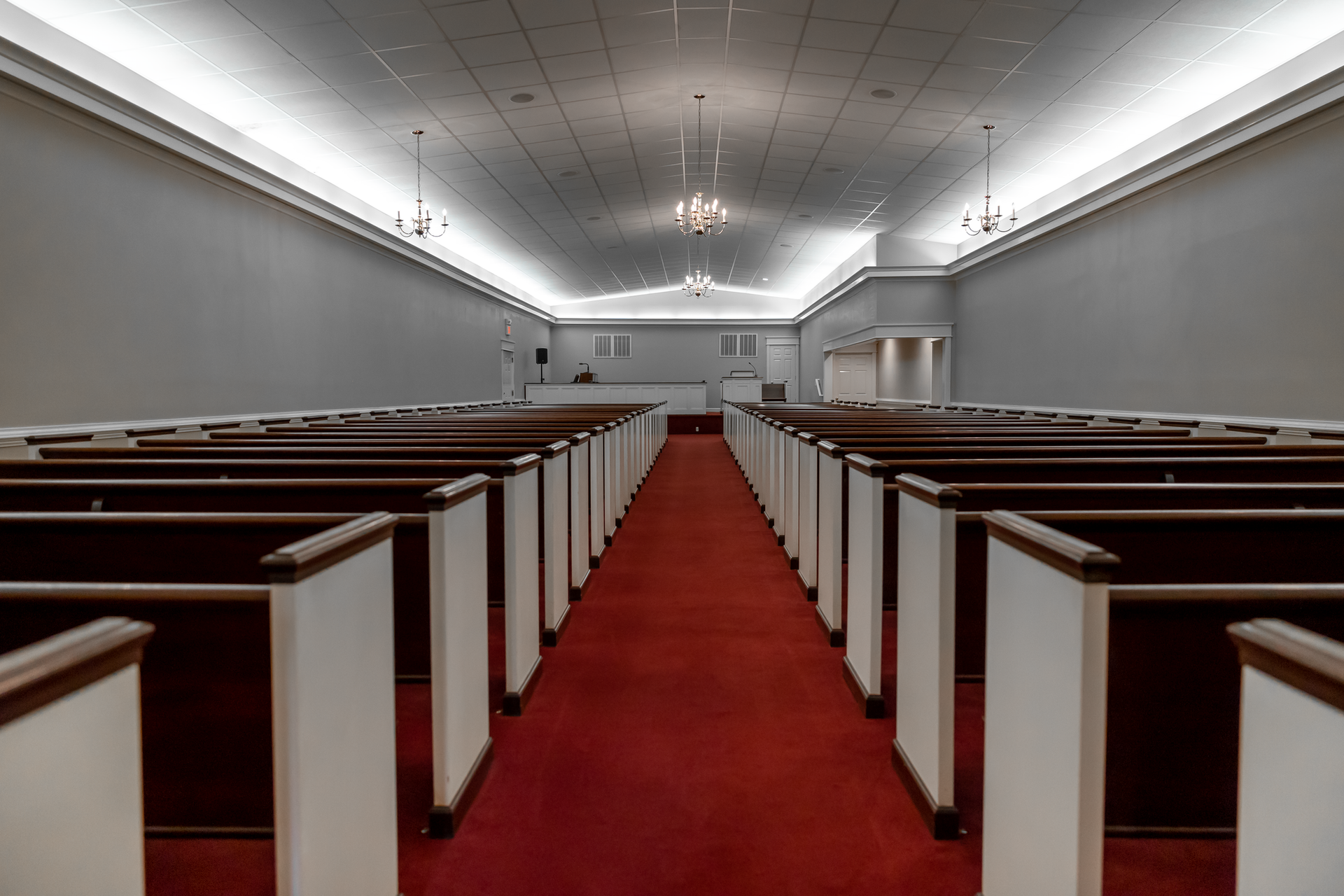 A view down the center aisle of an empty church sanctuary with rows of wooden pews and a red carpet leading to a stage.