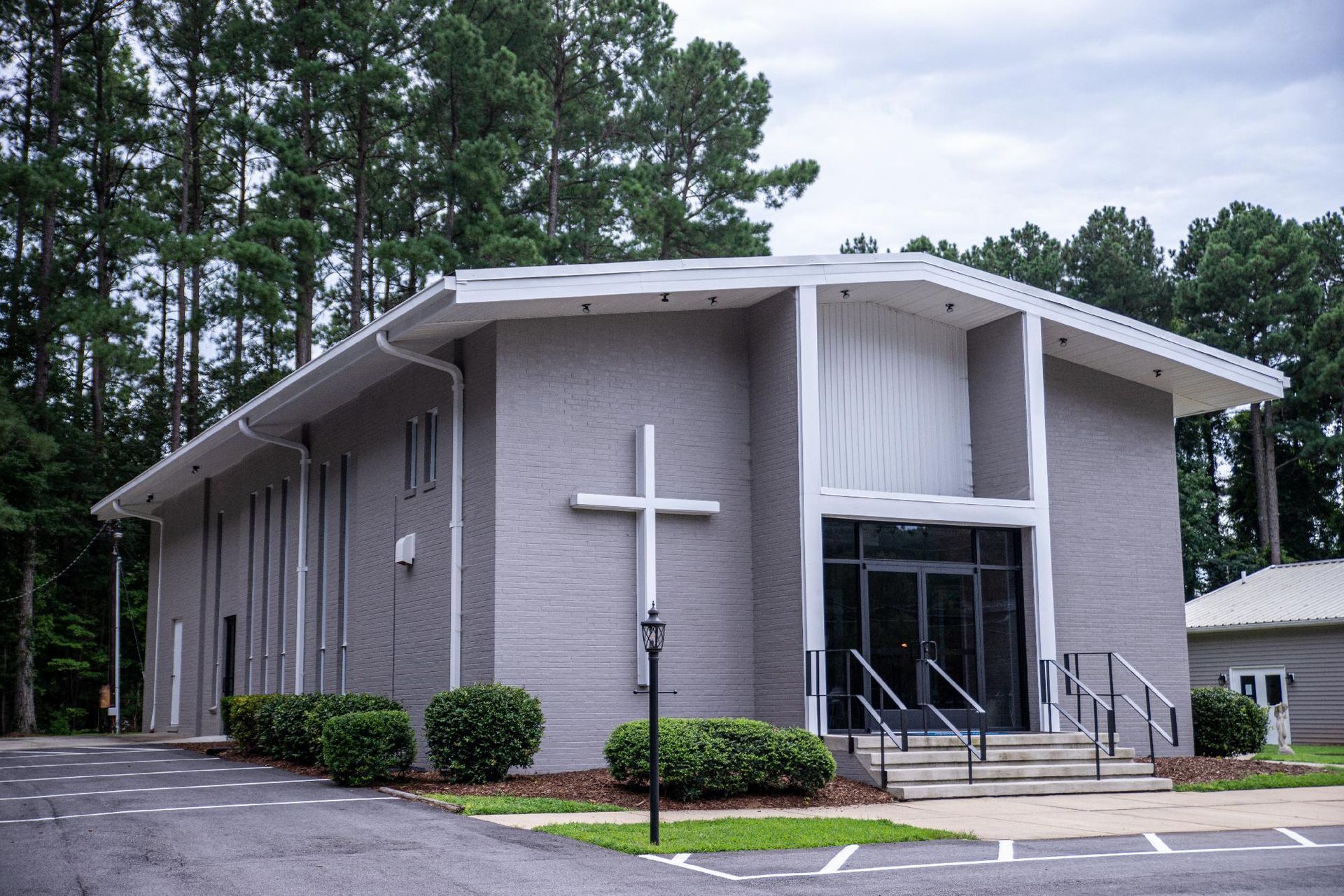 A grey, modern church building with a large white cross on the front facade, surrounded by trees and a parking lot.