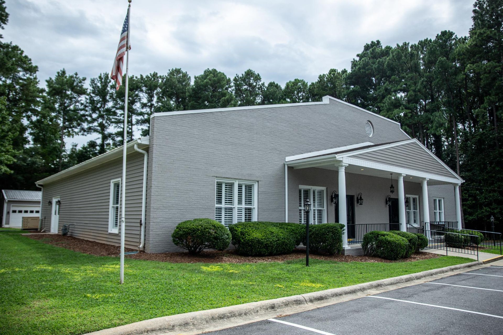 A single-story gray building with a porch and white pillars sits on a lawn beside a parking lot under a cloudy sky.