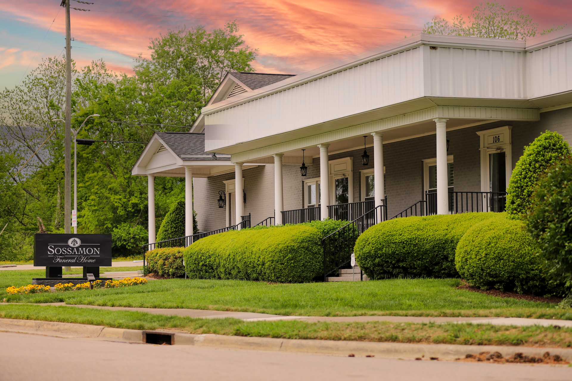 A gray building with a white porch and lush green bushes, featuring a sign reading "Seale Funeral Home" at sunset.