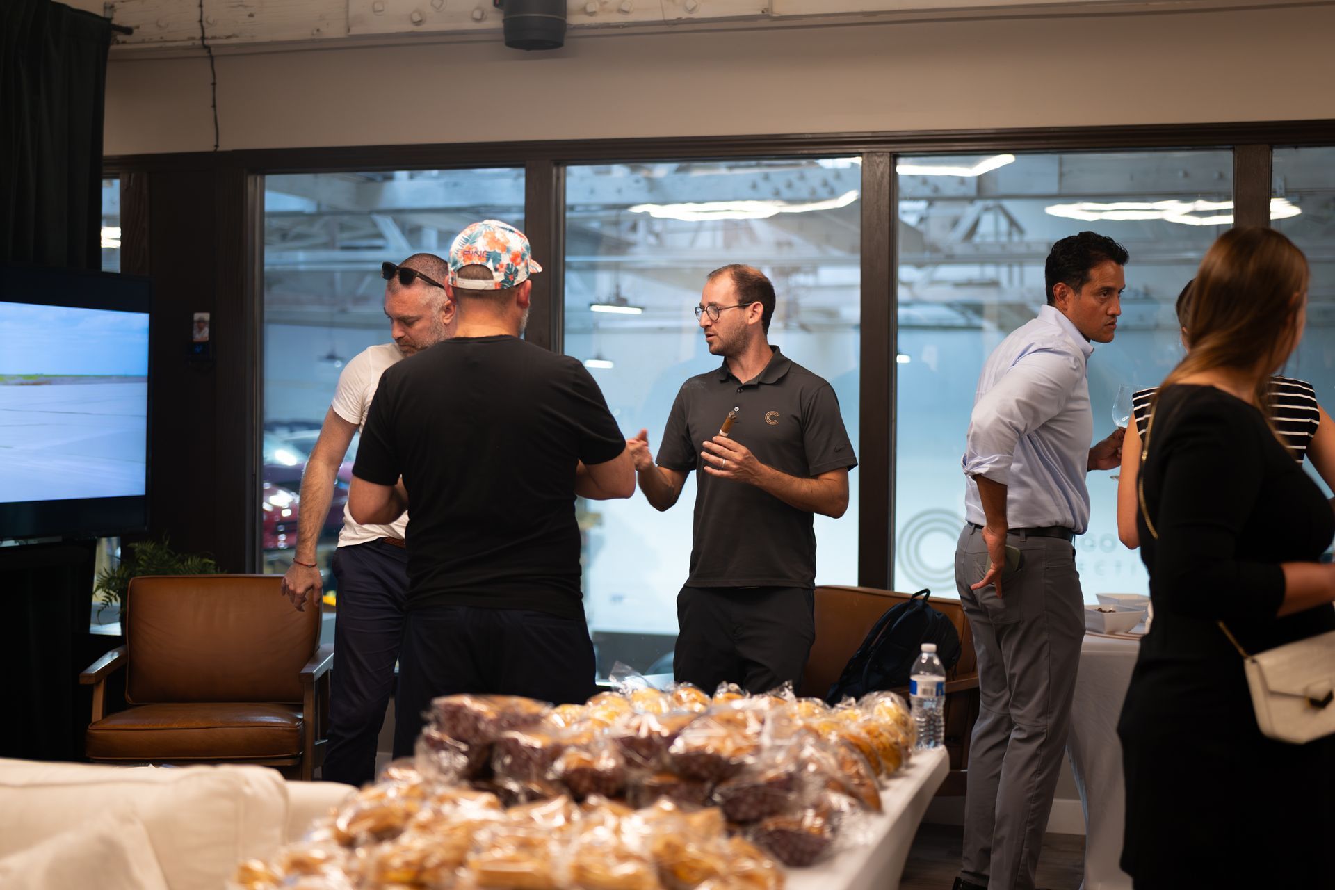 People socializing near a table of pastries. Man in gray shirt gestures. Window and TV in the background.