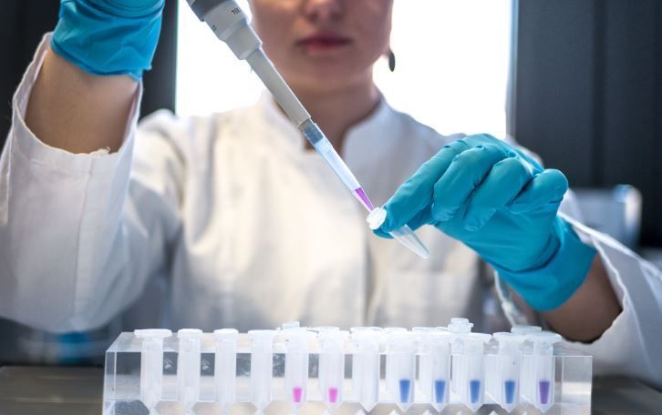 Una mujer con bata de laboratorio y guantes azules usa una pipeta para verter líquido en un tubo de ensayo.
