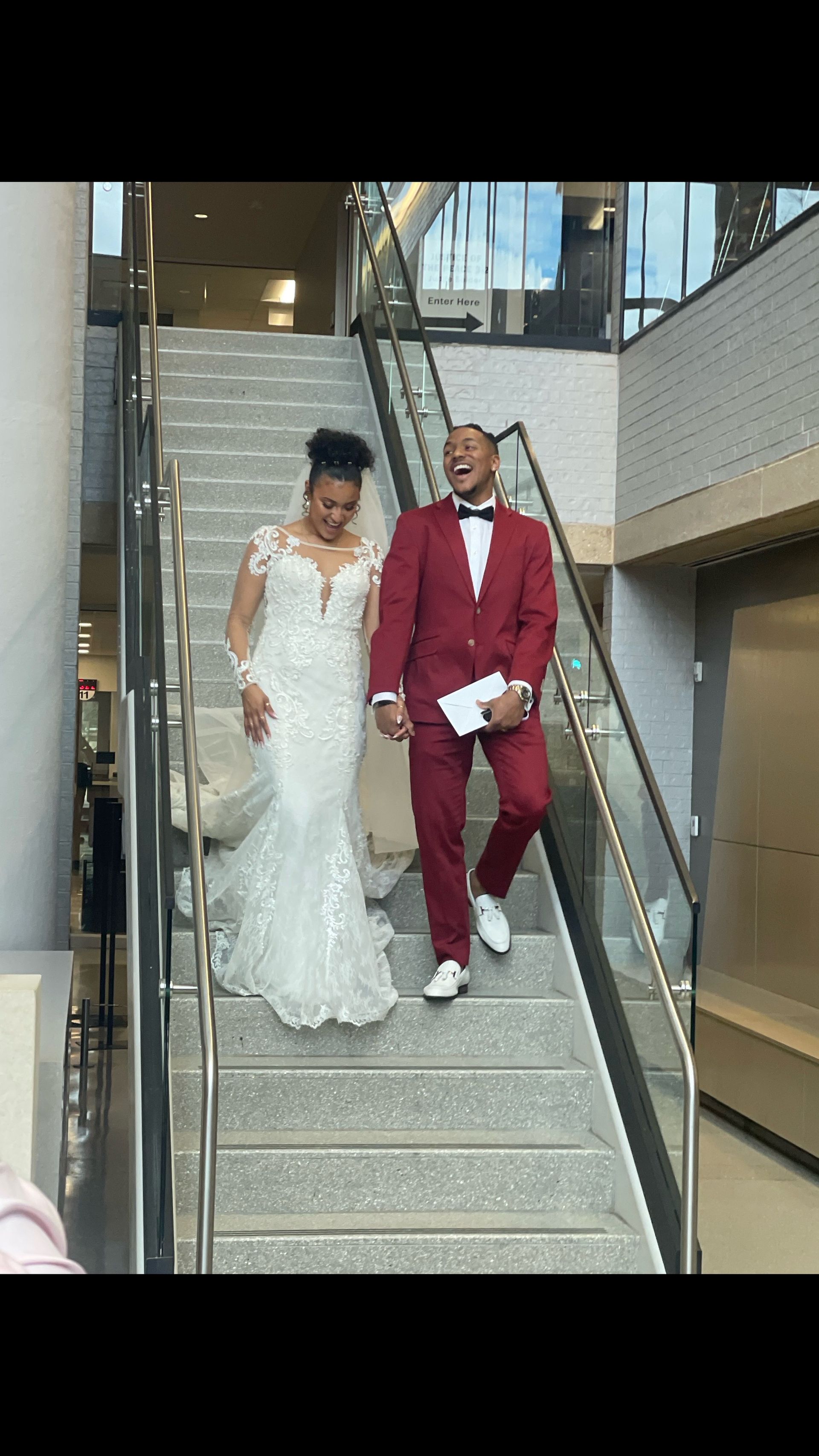 A bride in a white lace gown and a groom in a red suit walk down a staircase, both laughing.