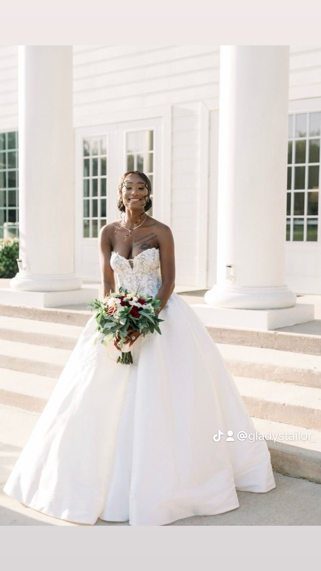 A smiling bride in a strapless white ballgown holds a floral bouquet while standing on steps between two white pillars.
