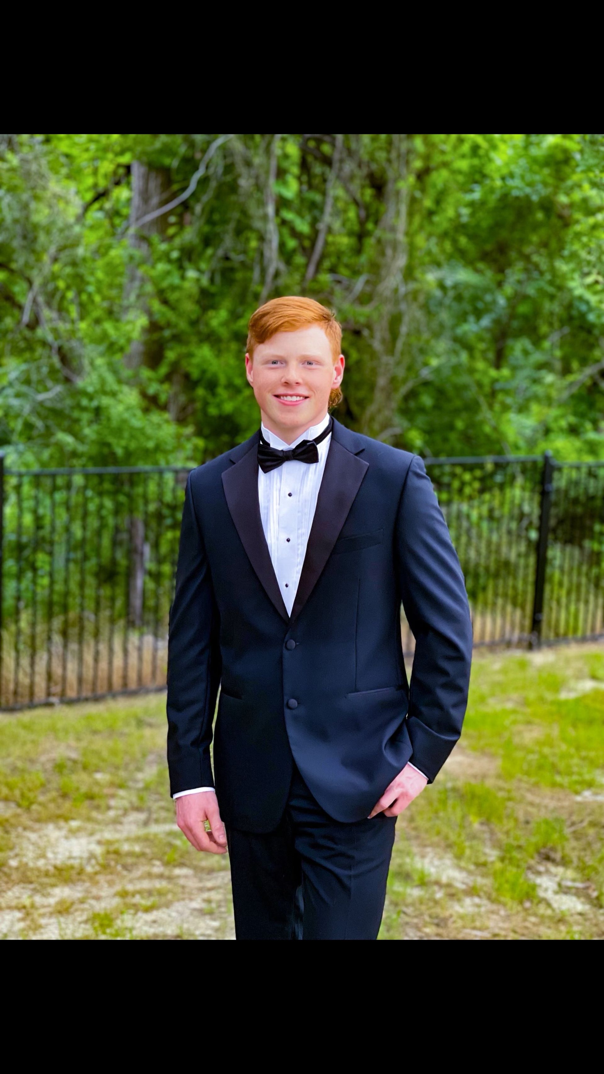 A young man in a tuxedo and bow tie is standing in front of a fence.