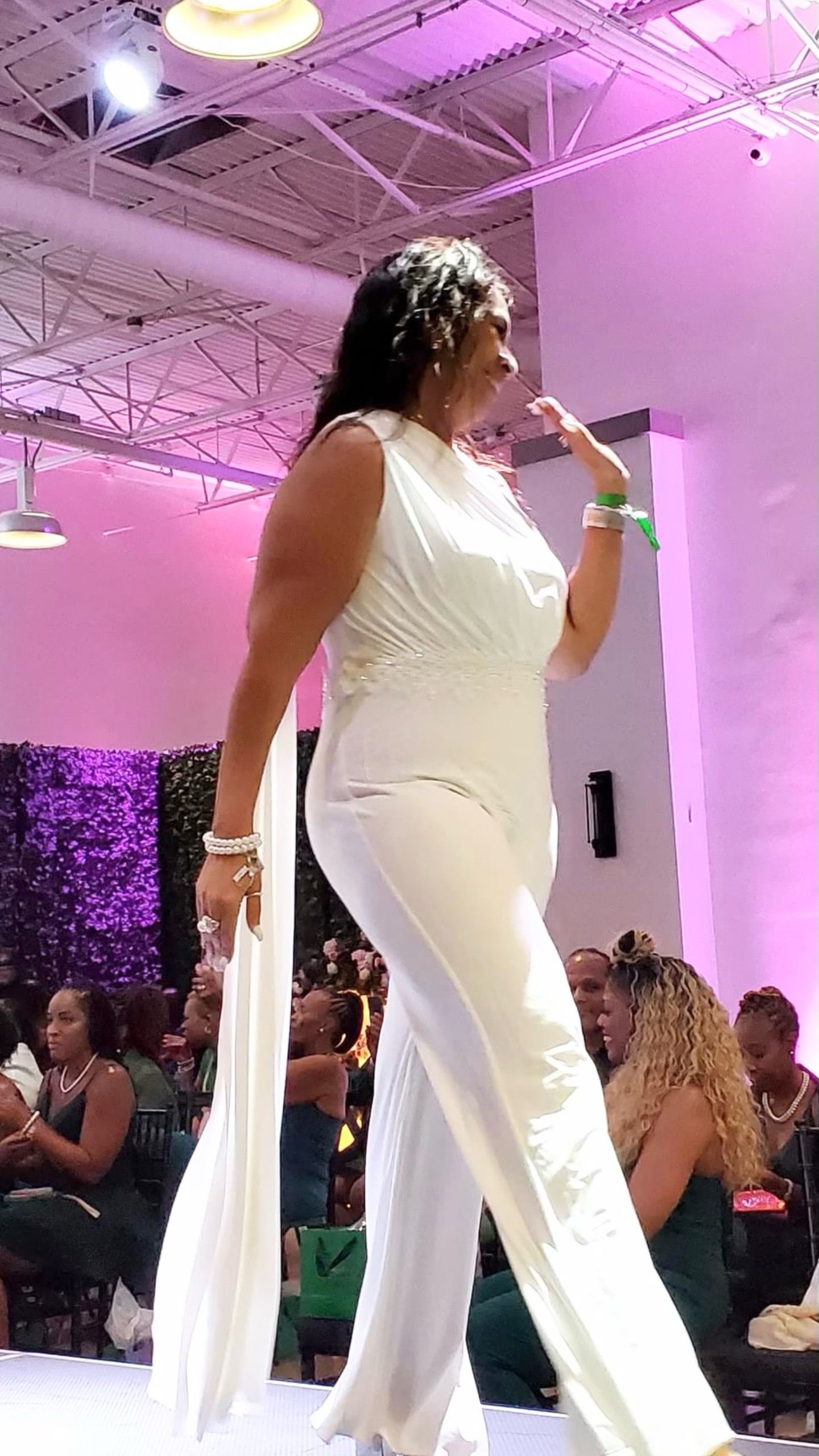 A person in a white jumpsuit with a flowing train walks along a runway during a fashion show with an audience nearby.
