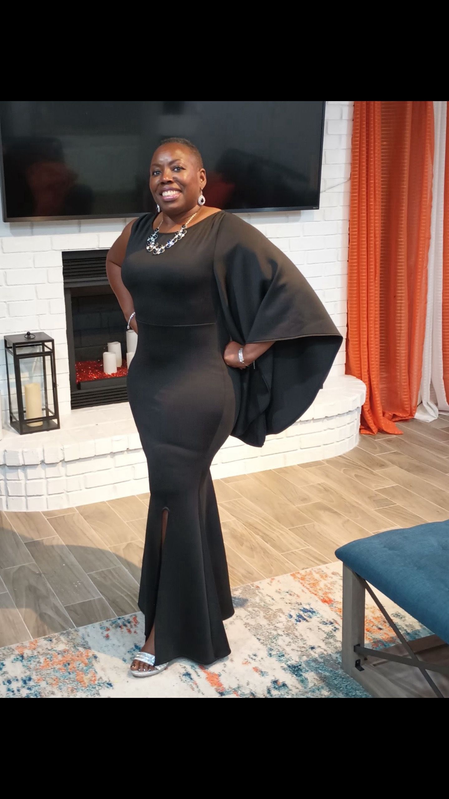 A woman in a black dress is standing in a living room in front of a fireplace.