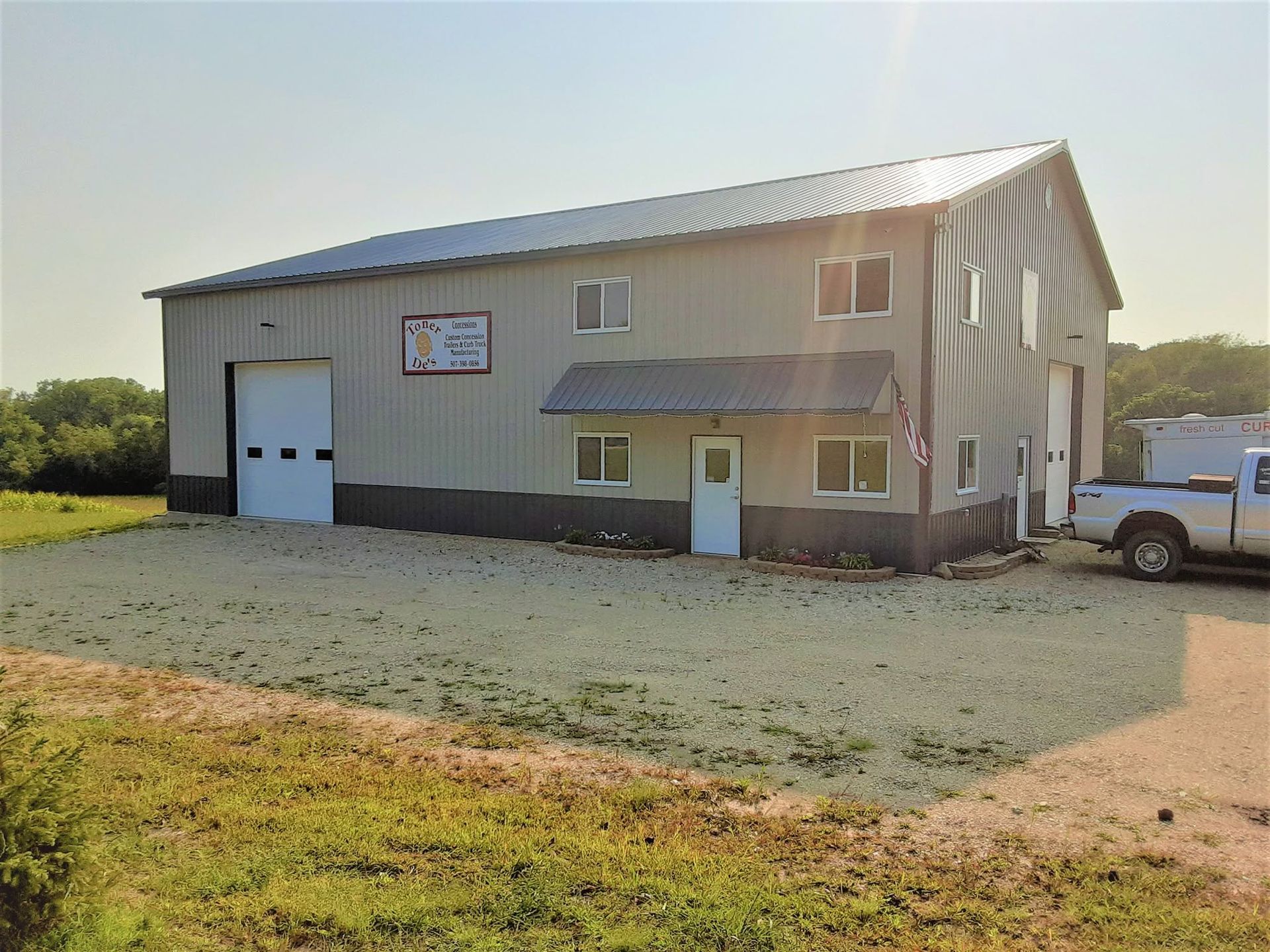 Metal workshop building with a garage door, white truck beside it, and gravel driveway in a rural setting