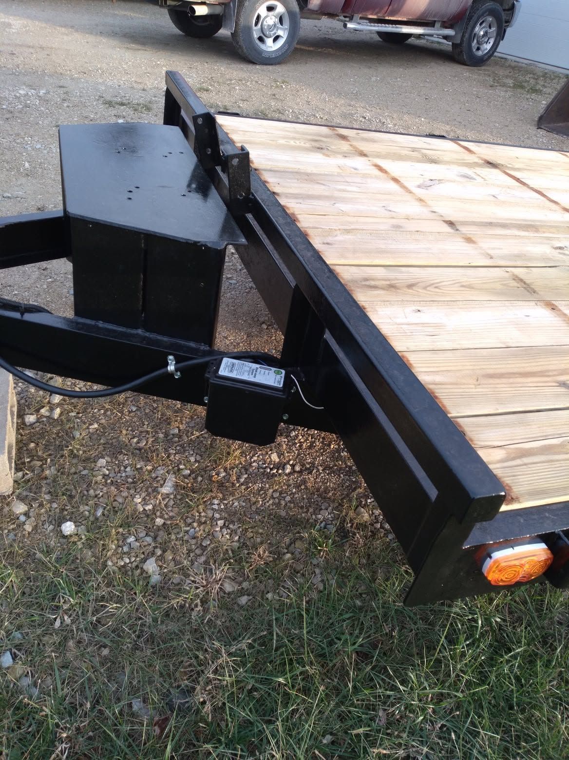 Black utility trailer with wood deck, hitch, and orange side reflector on gravel and grass.
