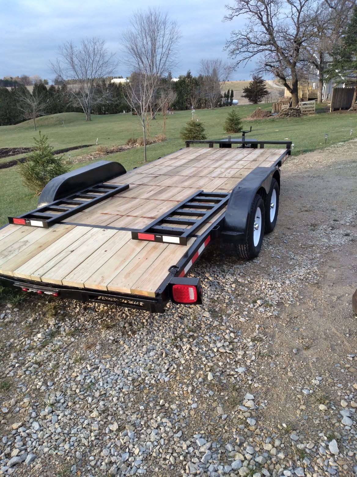 Empty flatbed trailer parked on gravel in a grassy yard, with ramps and a spare tire.
