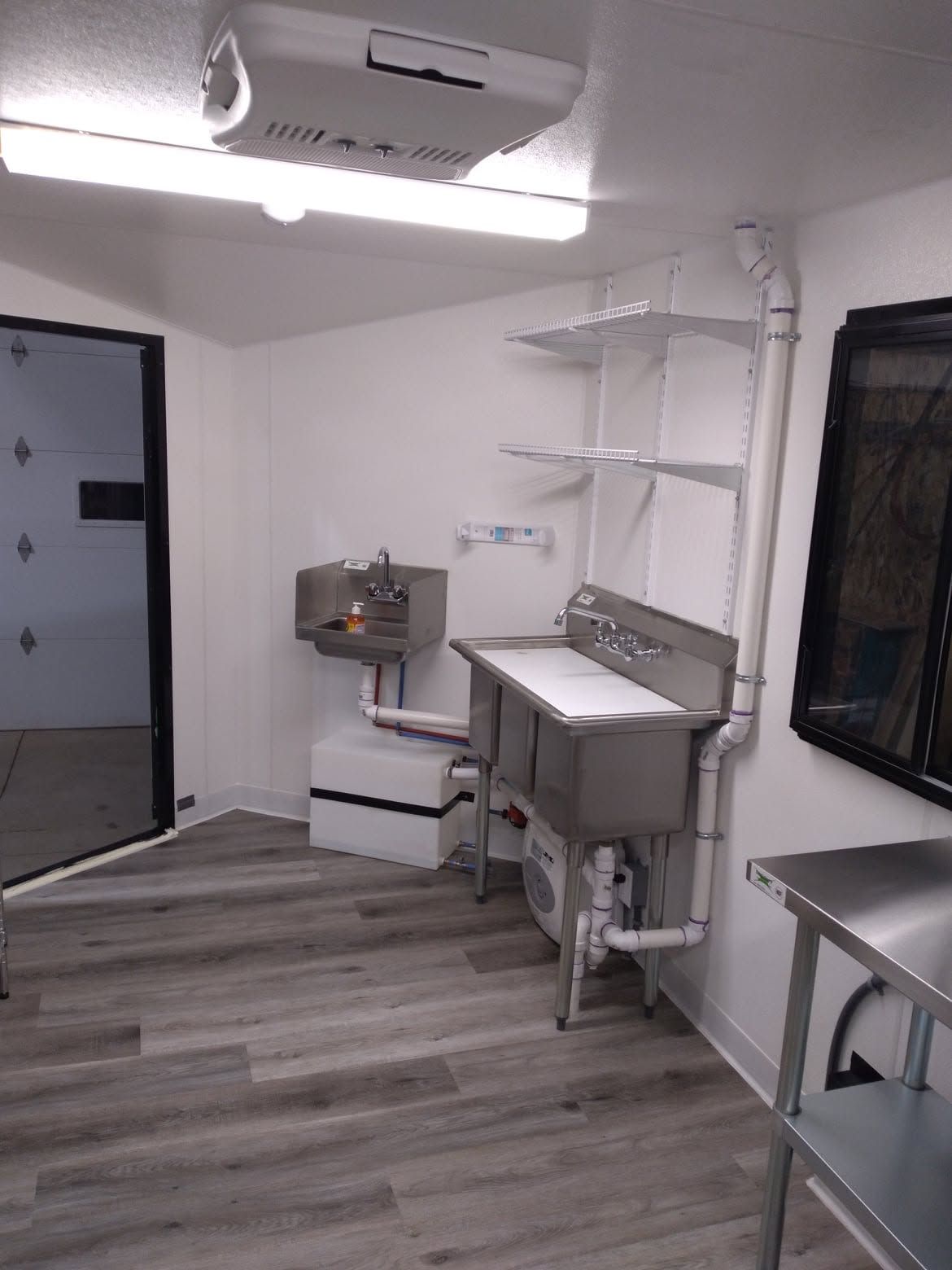 Small white utility room with sink, shelves, gray wood floor, and wall-mounted light fixtures.