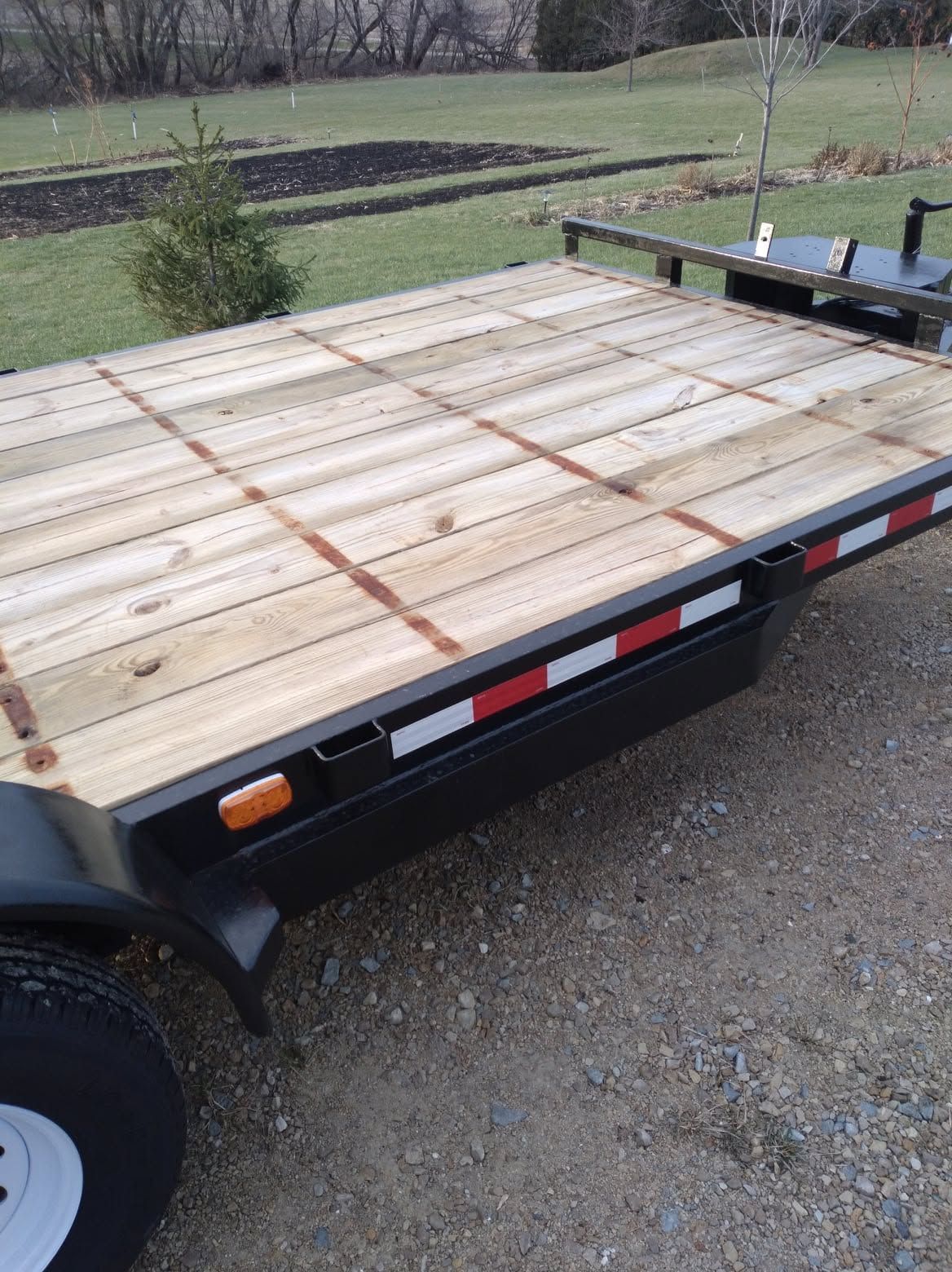 Flatbed utility trailer with a wooden deck parked on gravel in a rural setting.