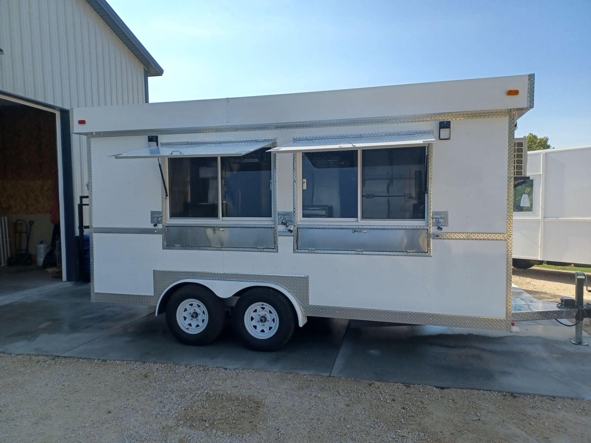 White double-axle food trailer with service windows parked beside a building on a sunny day