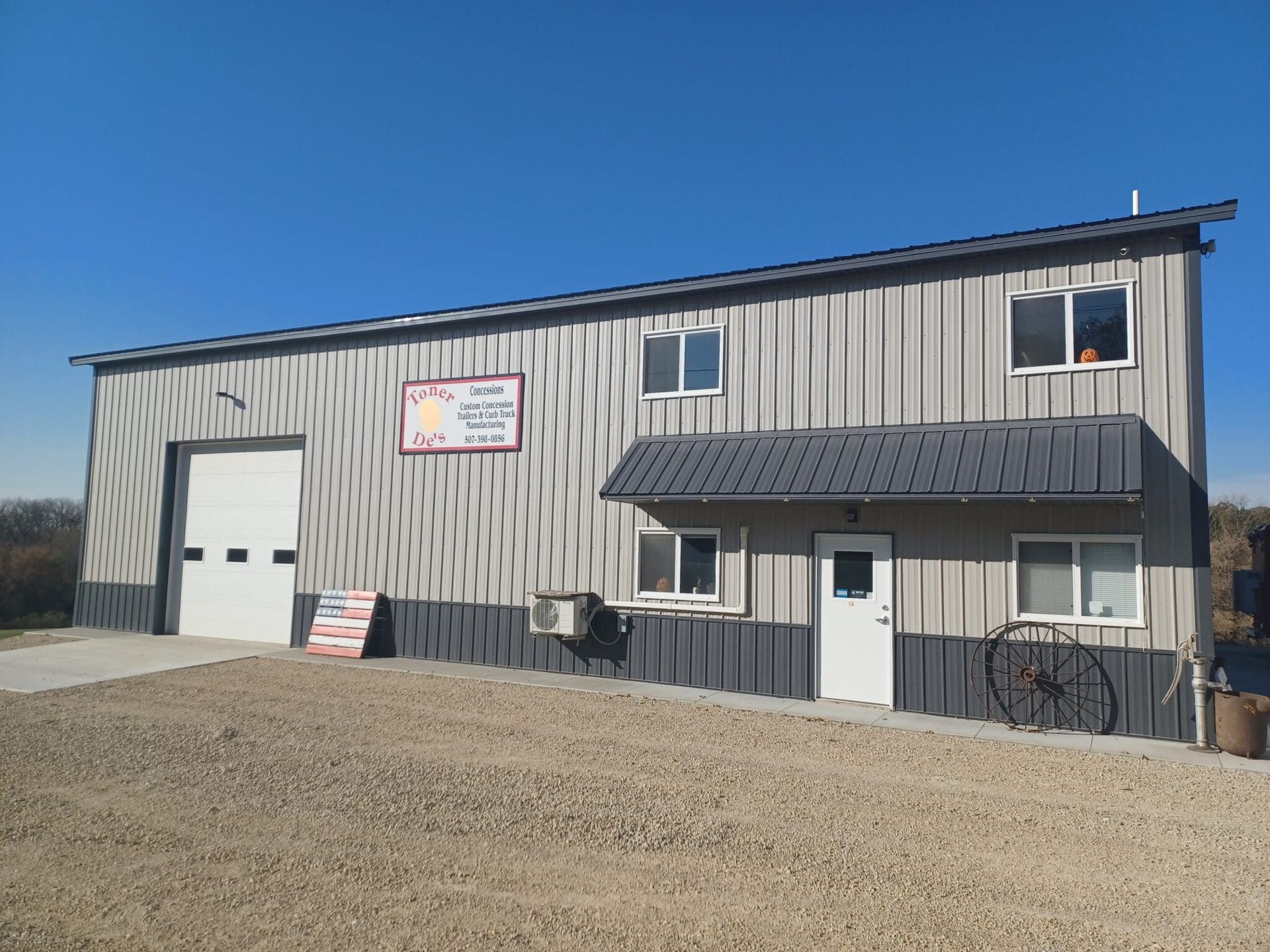Tan metal building with garage door, sign, and small porch on gravel lot under a clear sky