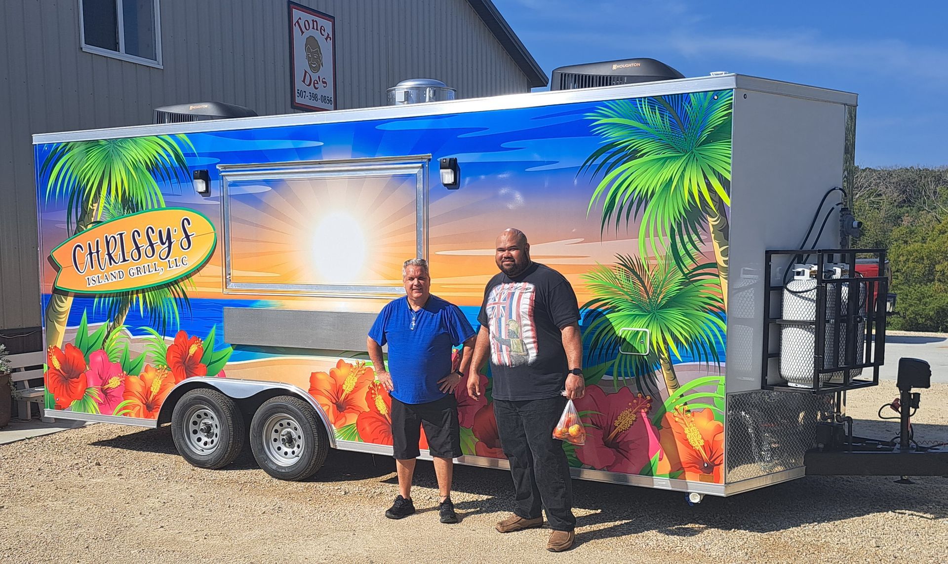 Two people stand beside a colorful tropical food trailer in a parking lot.