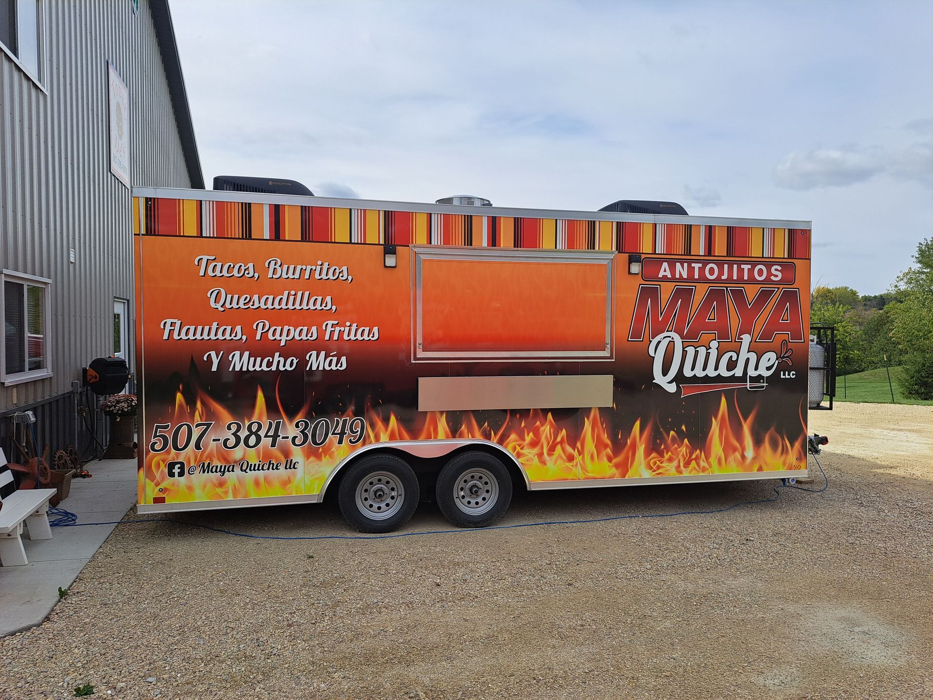 Bright orange food trailer with flame graphics and menu text parked beside a building.