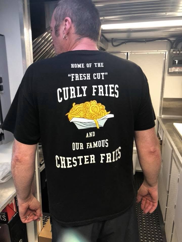 Man wearing a black T-shirt with a curly fries graphic and menu text on the back in a kitchen area